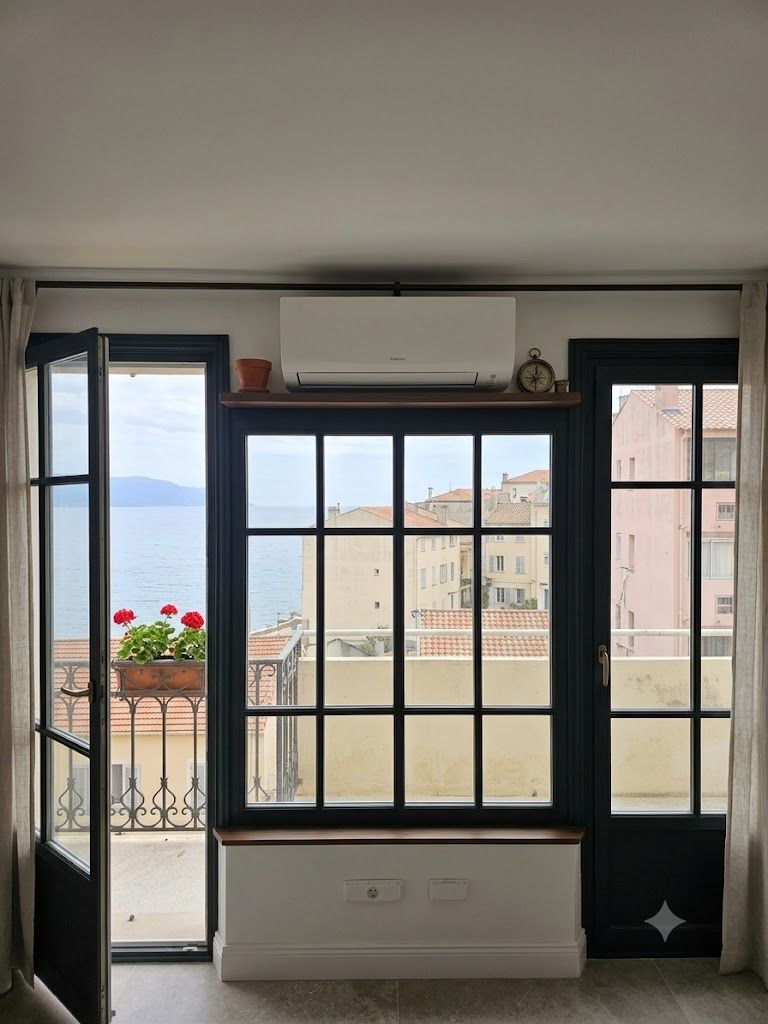 Chambre moderne avec de grandes fenêtres à cadre noir, vue depuis le balcon, radiateur blanc et fleurs rouges en pot.