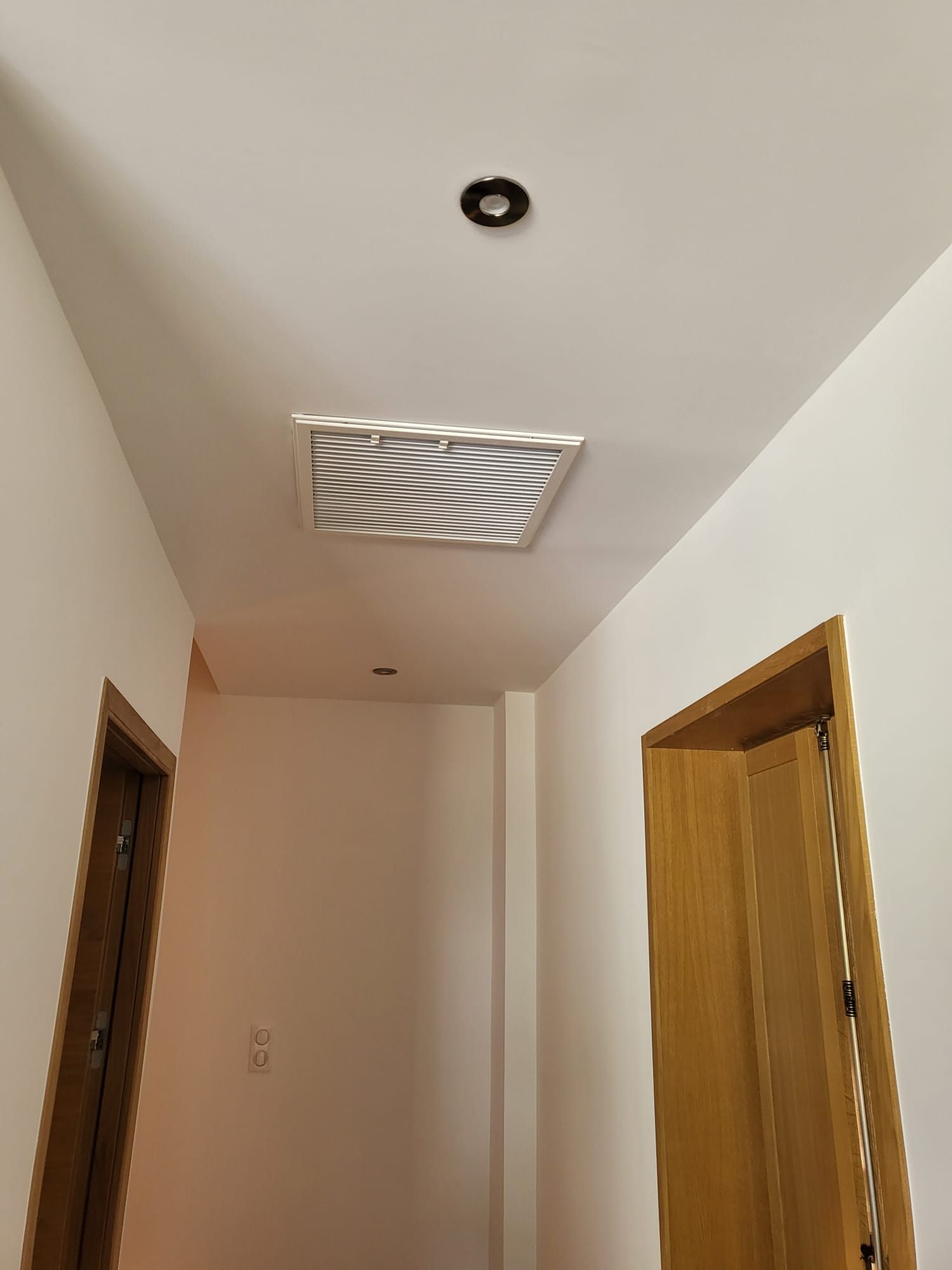 Couloir aux murs beiges, portes en bois, grille d'aération au plafond et spot encastré.