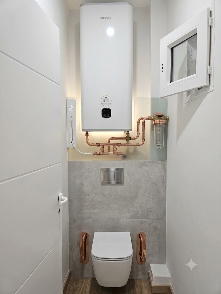 Petite salle de bains avec toilettes suspendues, chauffe-eau blanc, tuyaux de cuivre apparents et accents de carrelage gris.