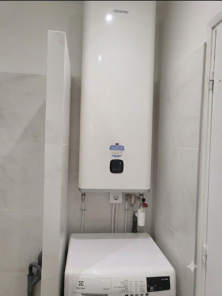 Chauffe-eau mural blanc avec tuyaux et vannes apparents dans une petite buanderie.