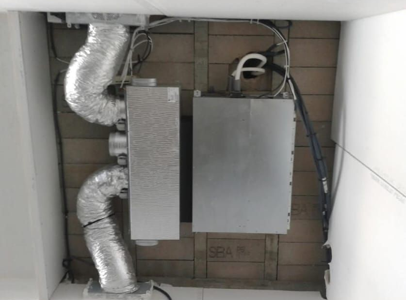 Unité de climatisation et conduits installés dans un local technique, avec grilles d'aération argentées et équipements métalliques contre un mur de briques