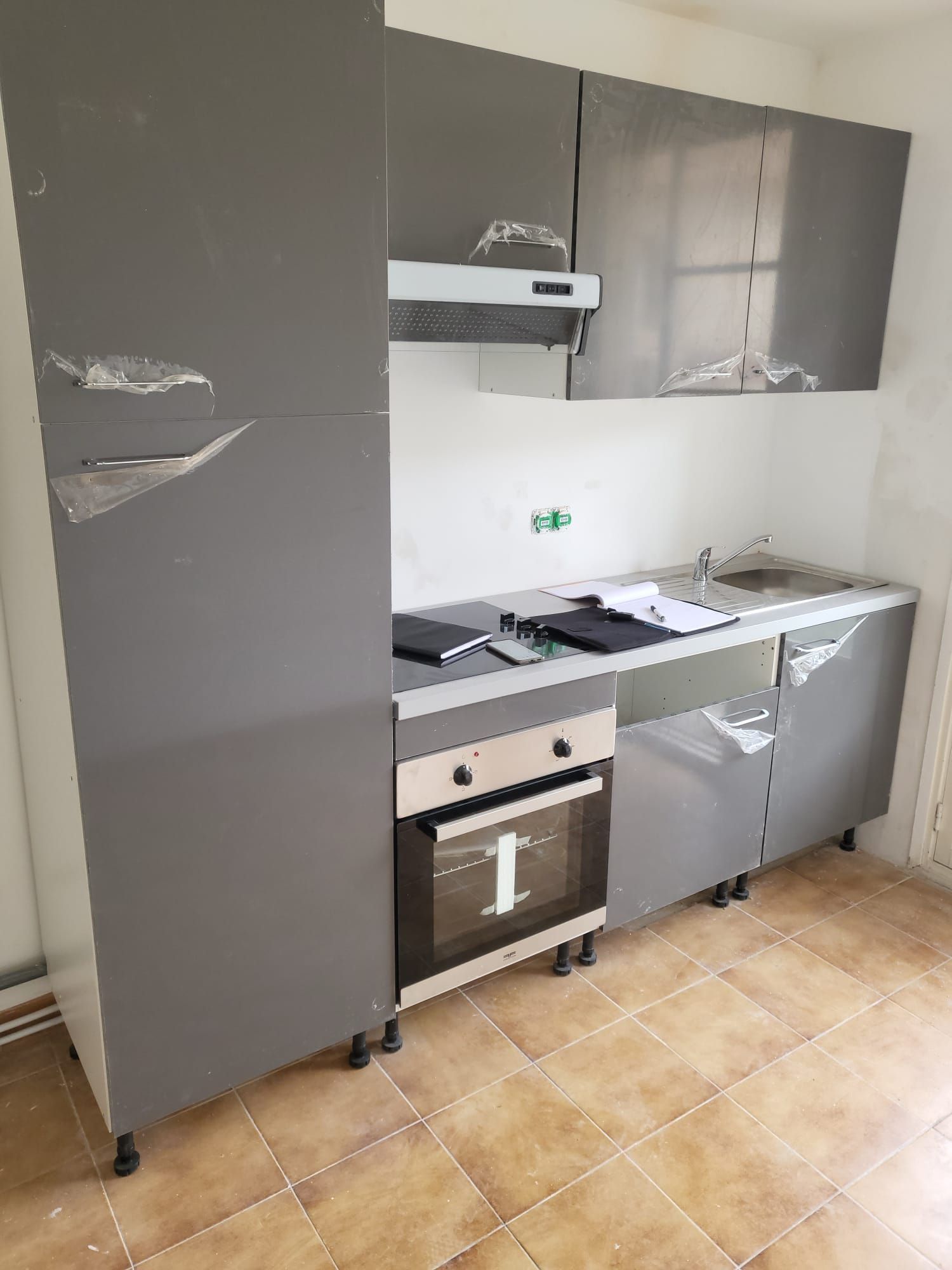 Petite cuisine avec placards gris, plan de travail blanc, cuisinière, évier et sol carrelé.
