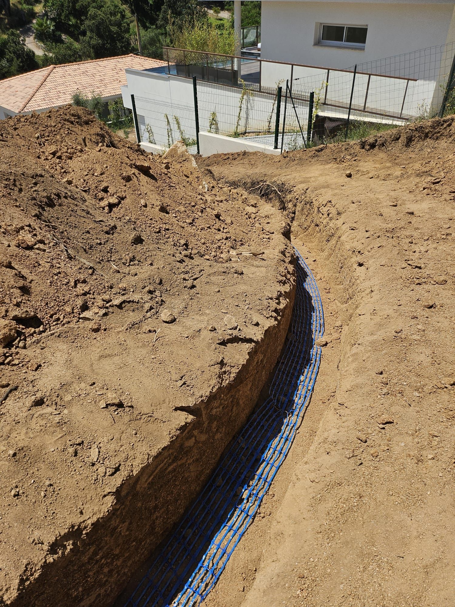 Tranchée incurvée tapissée d'un filet bleu sur un flanc de colline terreuse, à côté d'une maison blanche et d'une clôture.