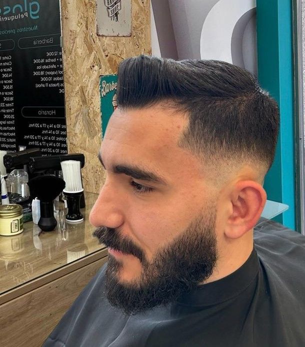 Hombre con un degradado marcado y barba completa, sentado en la silla de la barbería después de un corte recién hecho