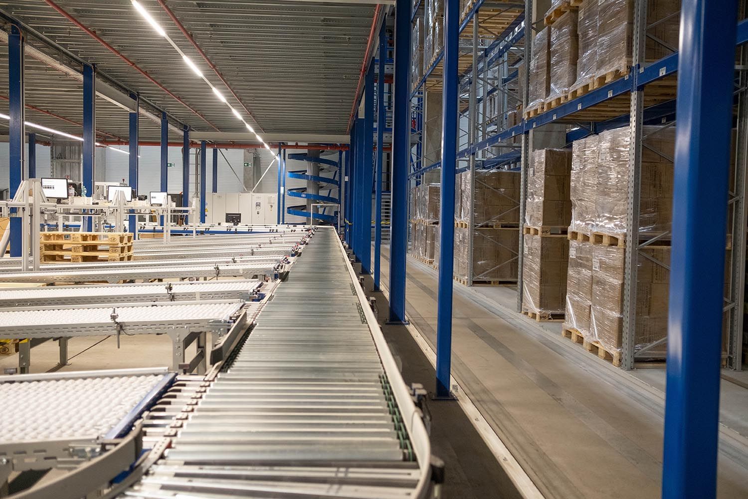 Lagerinnenraum mit Förderbandsystem und gestapelten Paletten. Blaue Stützen, Metall und Karton sichtbar.