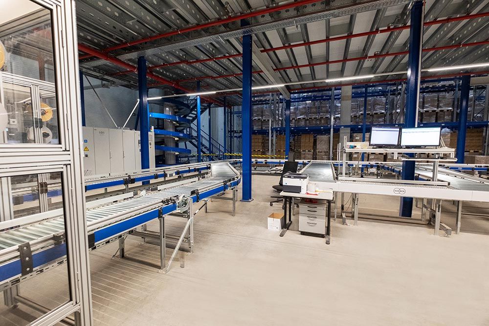 Förderbänder und Maschinen in einem Lagerhaus mit zwei Computermonitoren und einer Treppe, die in eine zweite Ebene führt.