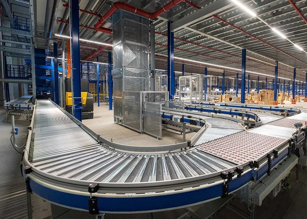 Förderbänder schlängeln sich durch das Innere einer Lagerhalle. Blau und Grau mit Deckenbeleuchtung.