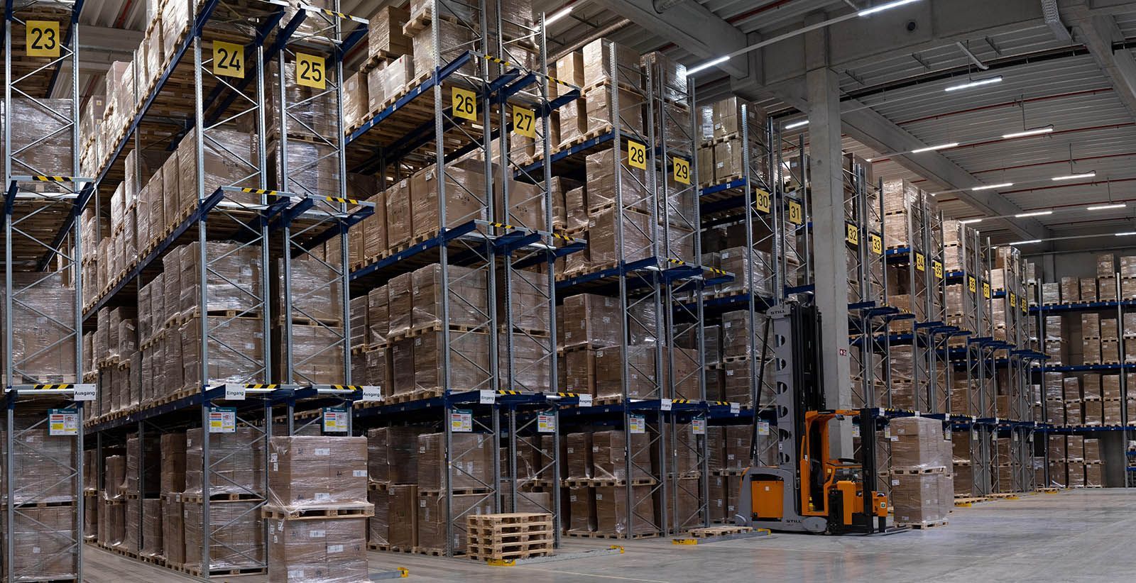 Lagerinnenraum mit hohen Regalen, in denen gepackte Kisten stehen, ein Gabelstapler ist in Betrieb.