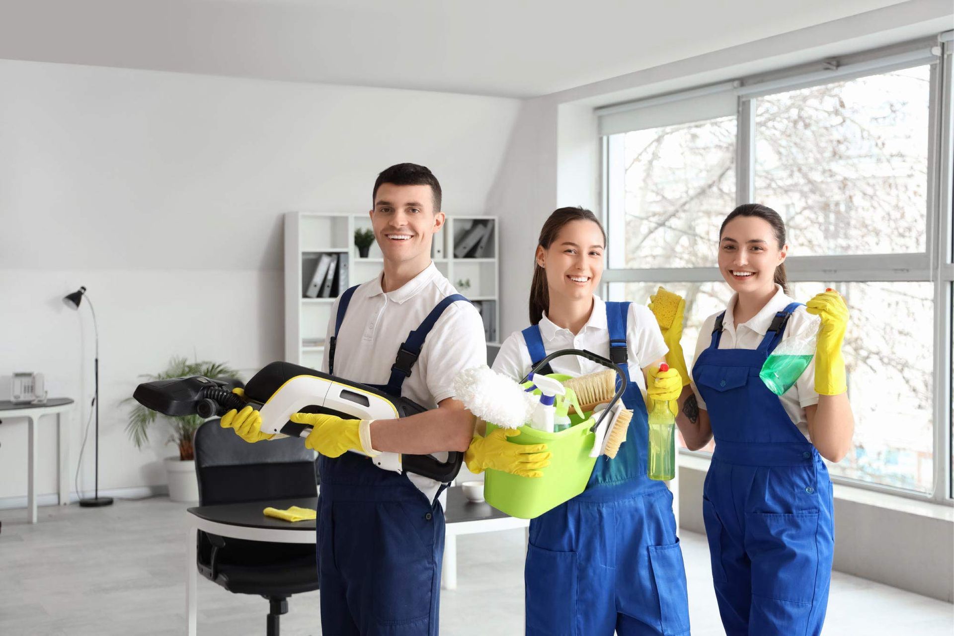Tres limpiadores sonrientes con monos azules y guantes amarillos en una oficina luminosa.