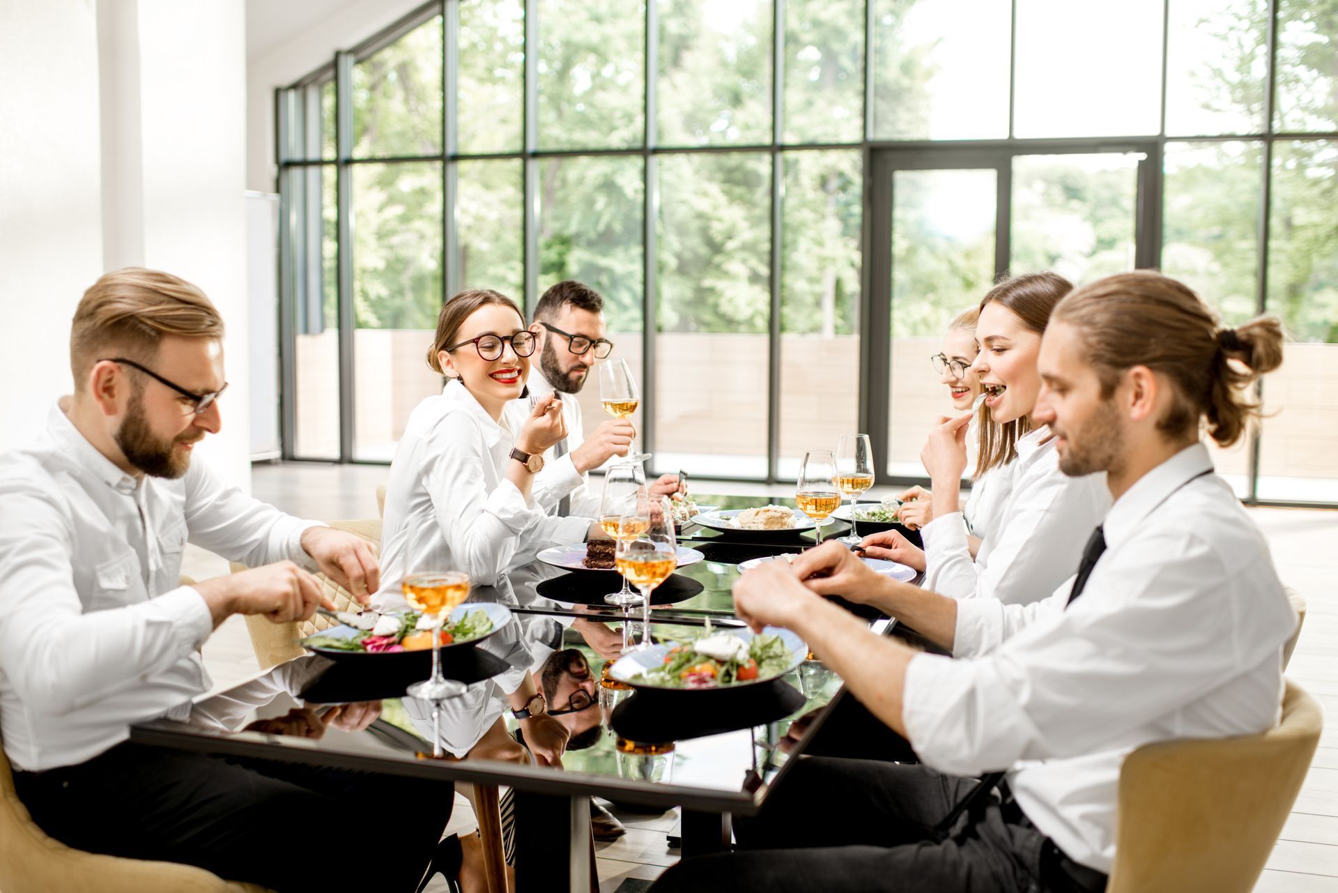Des personnes dînent à une table de conférence moderne avec de grandes fenêtres et de la verdure à l'extérieur.