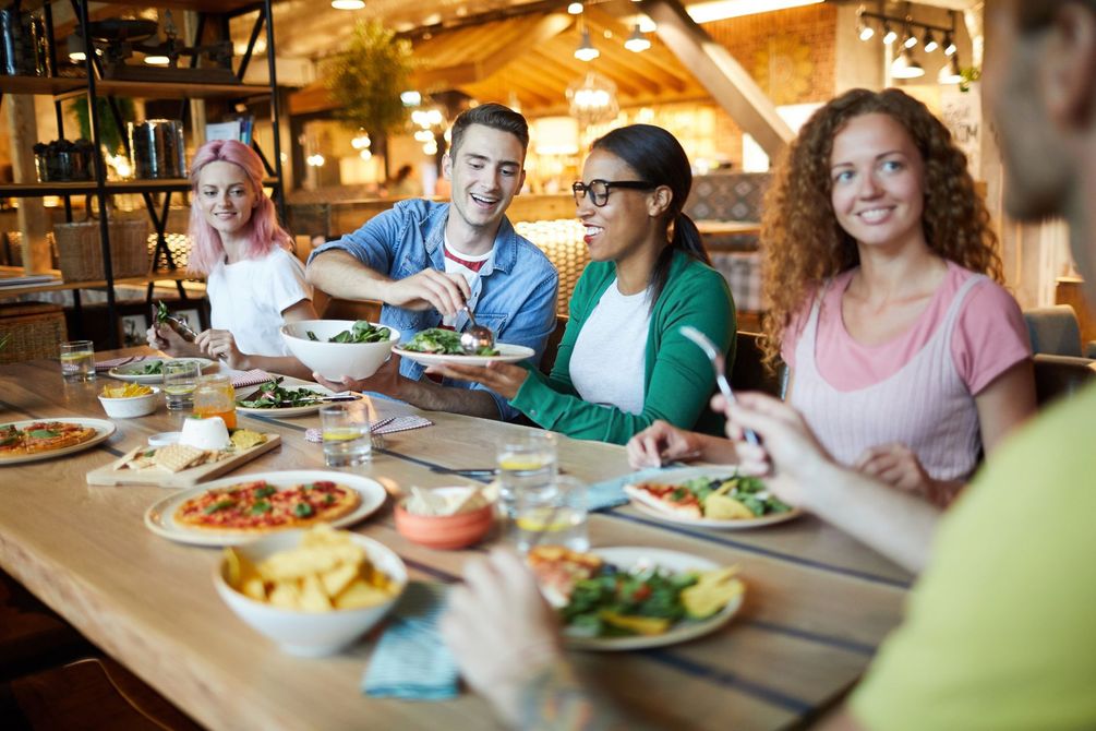 Des amis partagent un repas à une table de restaurant avec des pizzas, des salades et des boissons dans un cadre chaleureux et convivial.