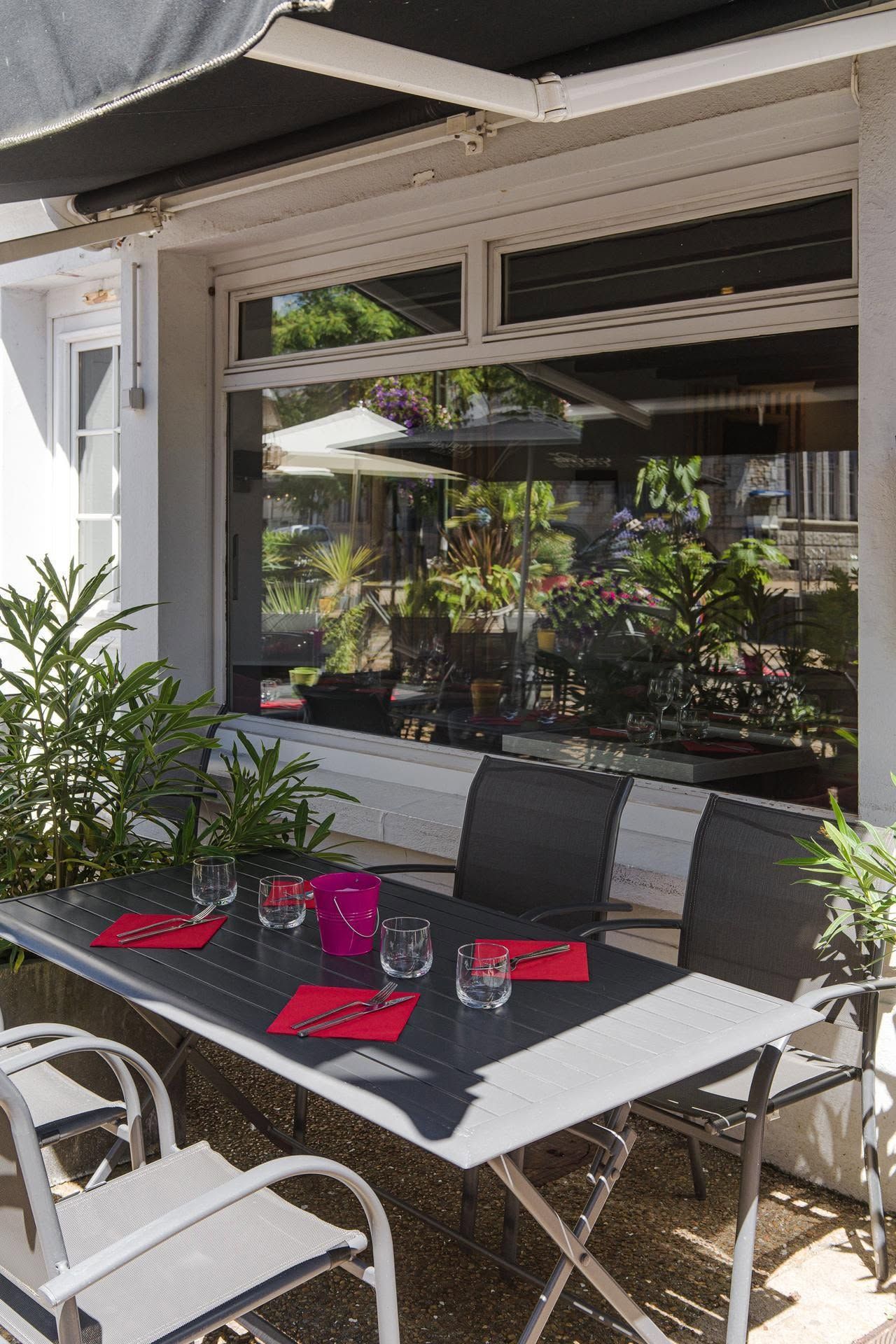 Table de café en terrasse avec sets de table et verres près d'une fenêtre et de plantes en pot.