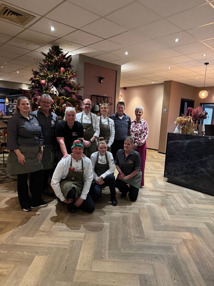 Medewerkers van Hotel Talens poseren in een hotellobby naast een versierde kerstboom en een open haard op de achtergrond.