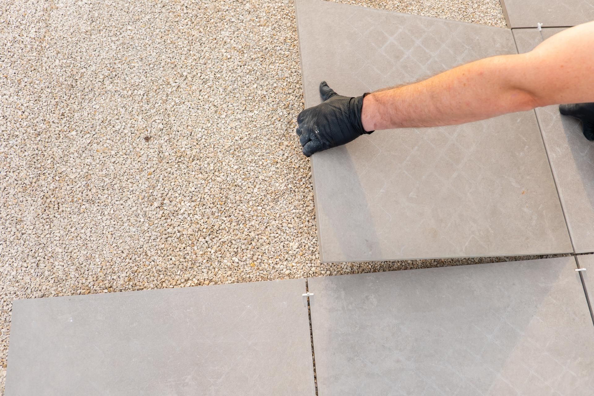 Eine behandschuhte Hand richtet eine graue Pflastersteinfliese neben einer gesprenkelten Kiesfläche aus.