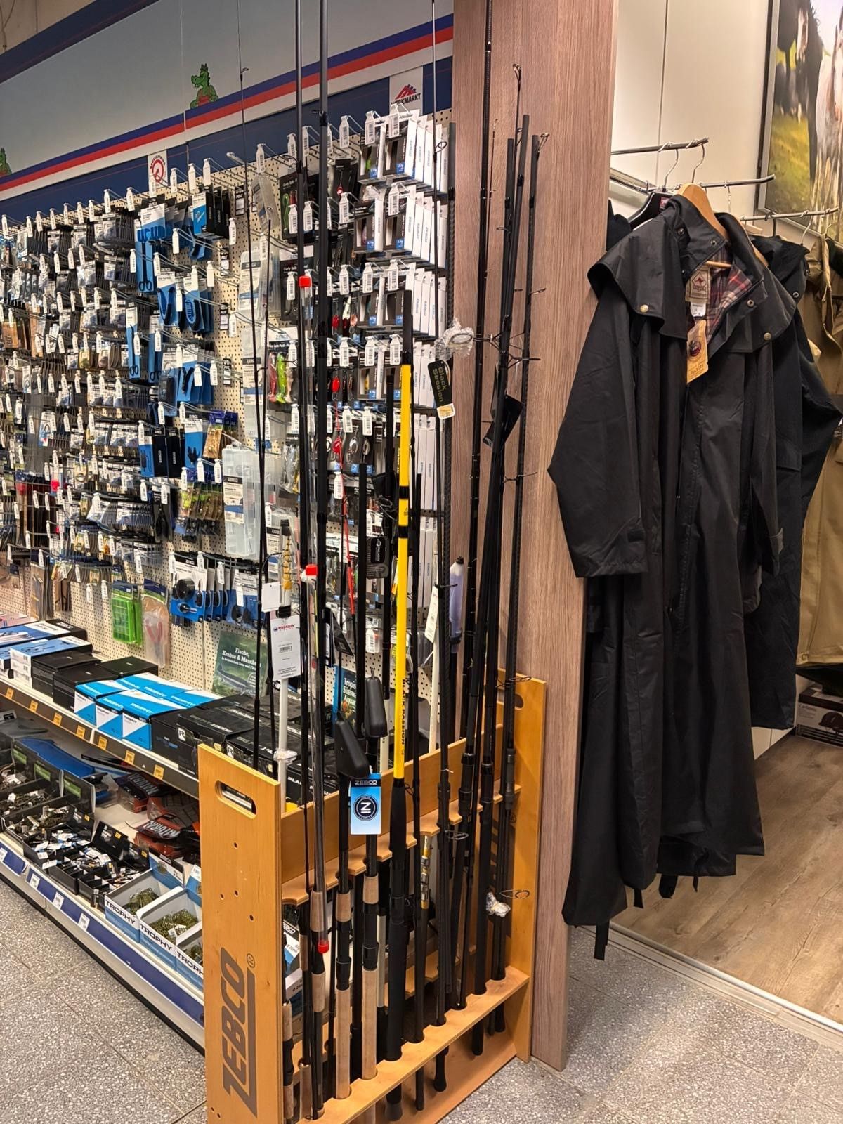 Angelruten und Angelzubehör Angelruten und Angelzubehör sind in einem Ladenregal neben hängenden schwarzen Jacken ausgestellt.