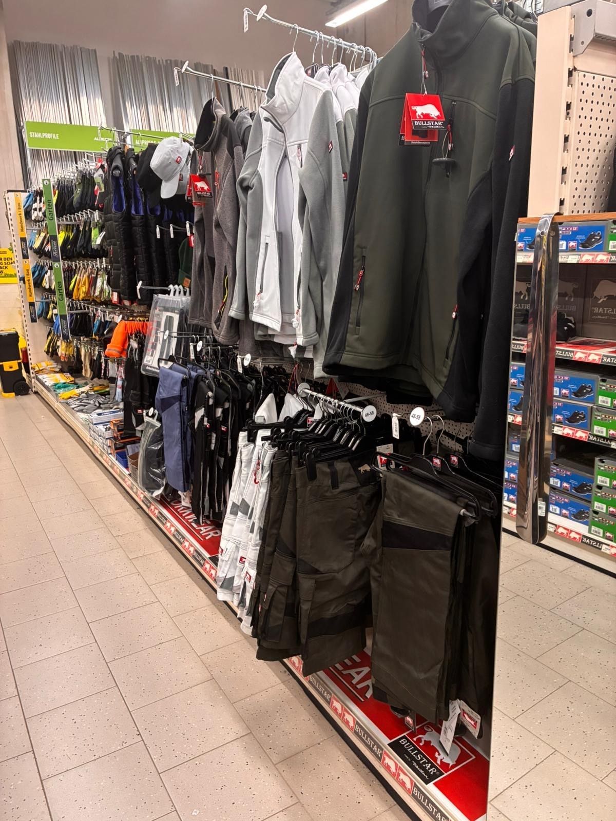 Gang in einem Bekleidungsgeschäft mit hängenden Jacken, Hosen und Accessoires an Kleiderständern.