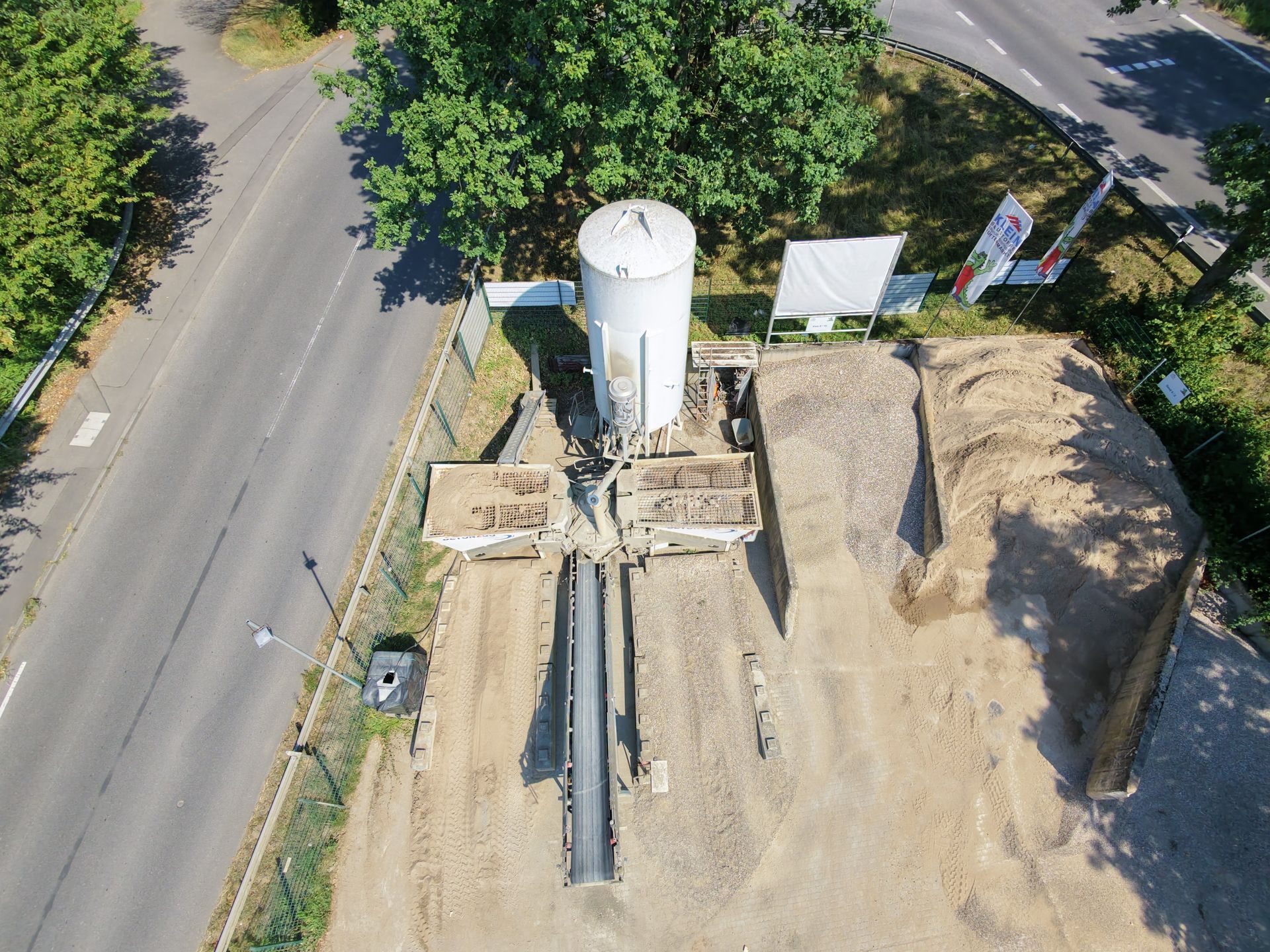Luftaufnahme einer Straßenbaustelle mit einem hohen weißen Silo, Erdhaufen und Baumaschinen neben einer Straße