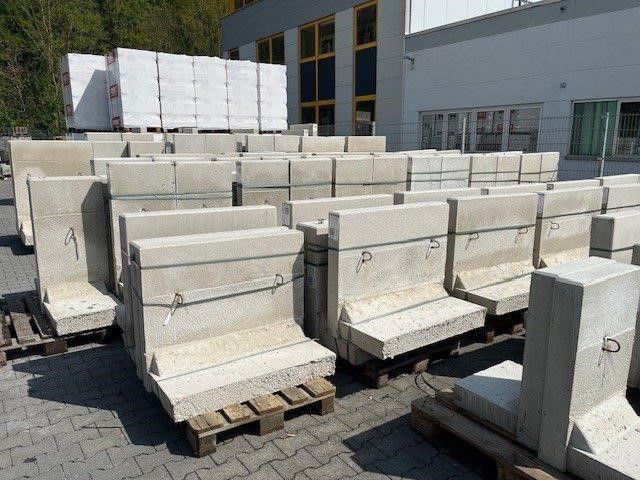 Gestapelte Betonbarrieren und -blöcke auf Paletten vor einem Industriegebäude.