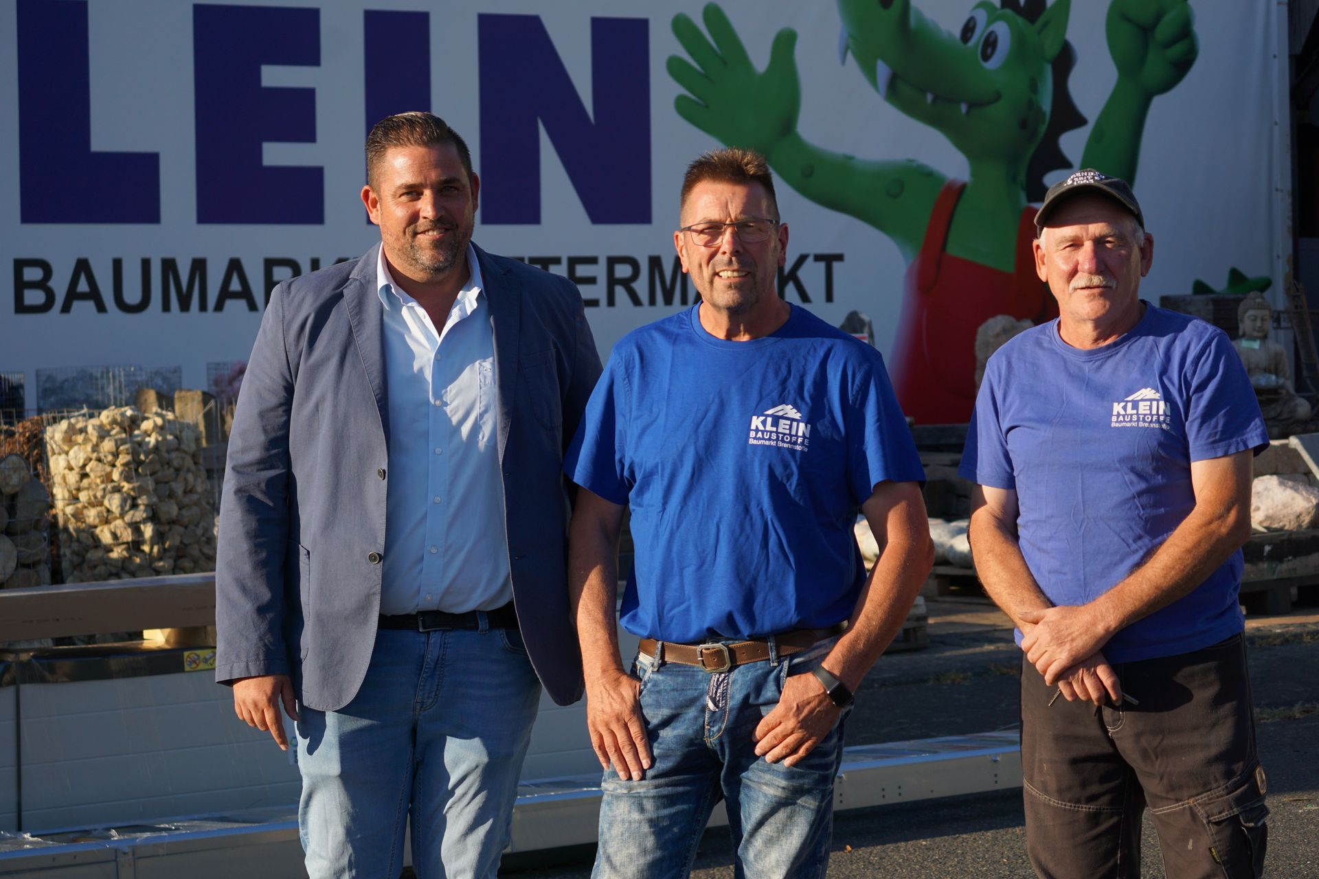 Drei Männer stehen lächelnd im Freien vor einem KEIN-Baumarkt-Schild. Zwei tragen blaue Hemden, einer ein Sakko