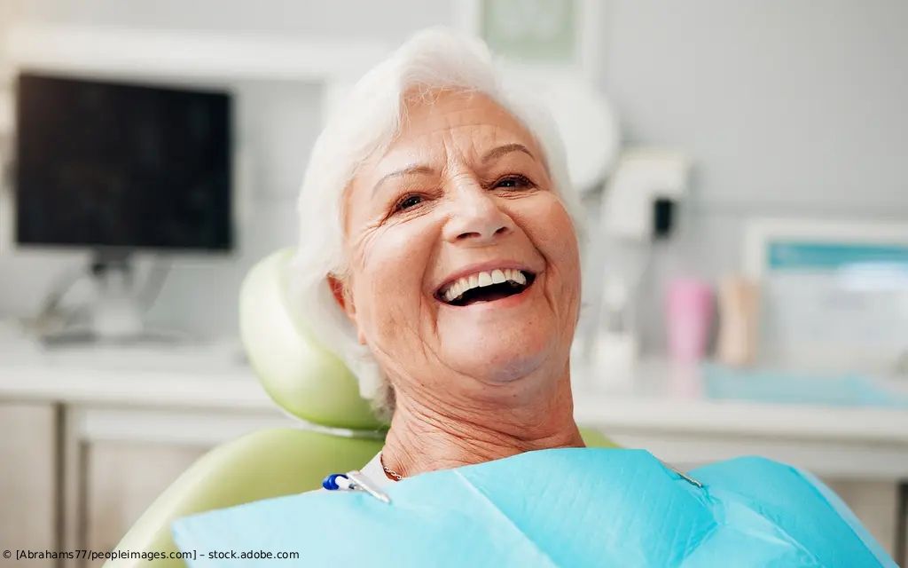 Ein lächelnder Patient mit blauem Lätzchen liegt in einem Zahnarztstuhl in einer hellen Klinik.