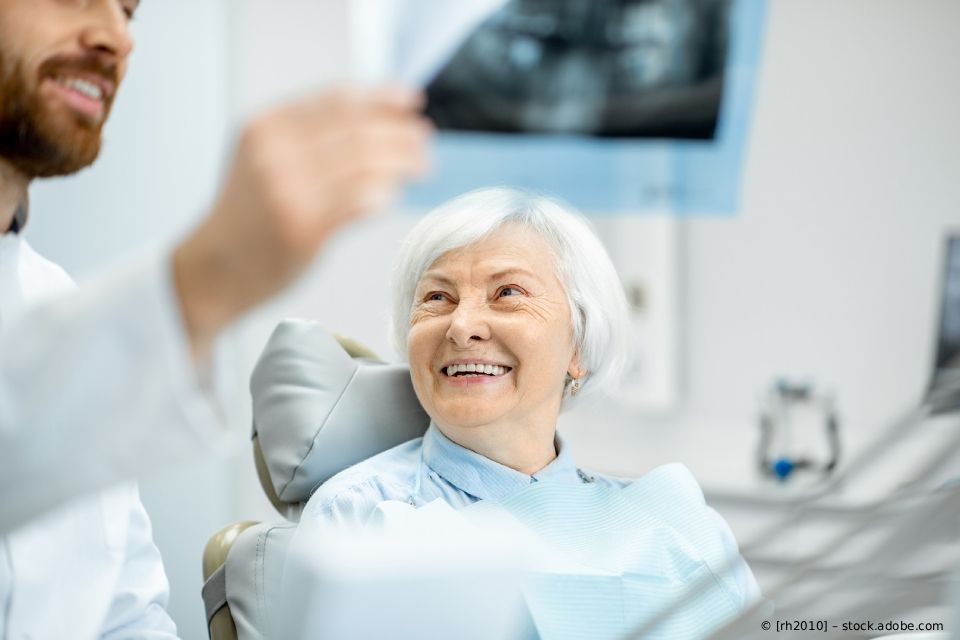 Ein lächelnder Patient sitzt auf einem Zahnarztstuhl, während der Zahnarzt ein Röntgenbild hochhält.