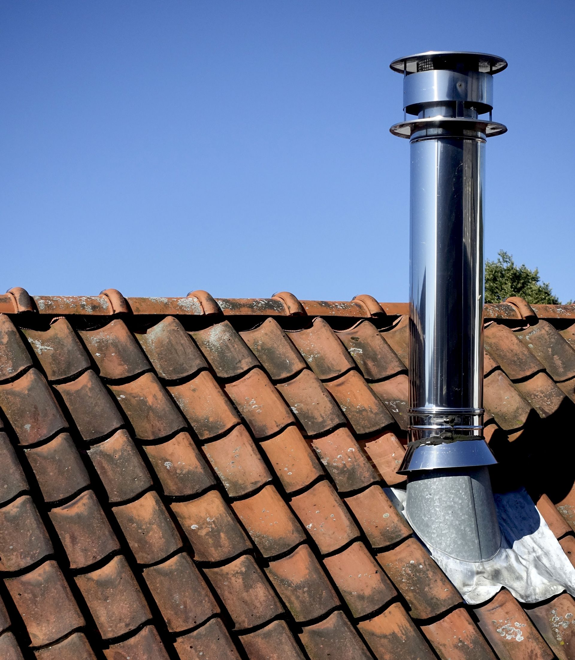 Cheminée en métal brillant sur un toit de tuiles rouges se détachant sur un ciel bleu limpide.