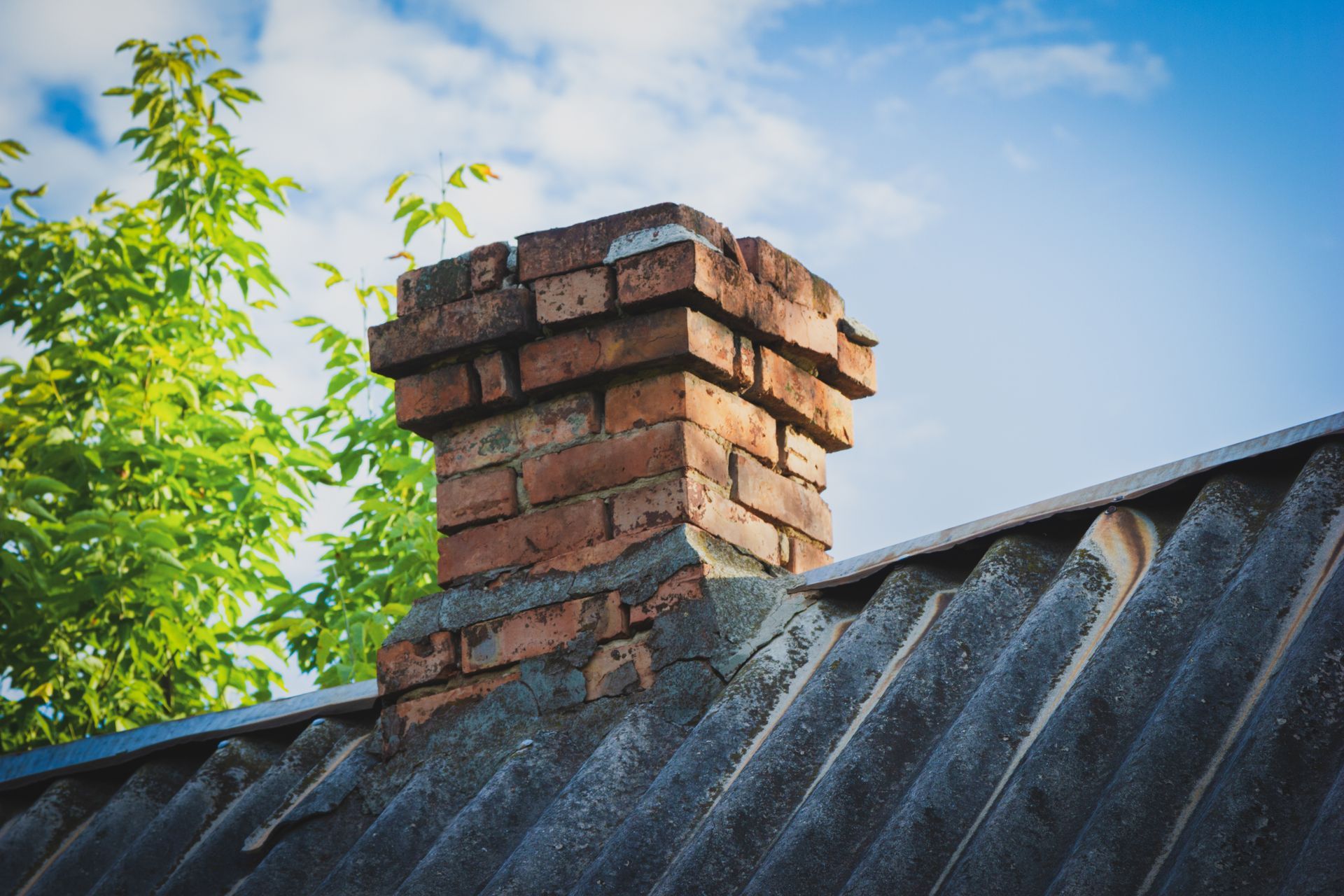 Cheminée en briques sur un toit de tôle ondulée grise, se détachant sur un ciel bleu parsemé de feuillage vert.