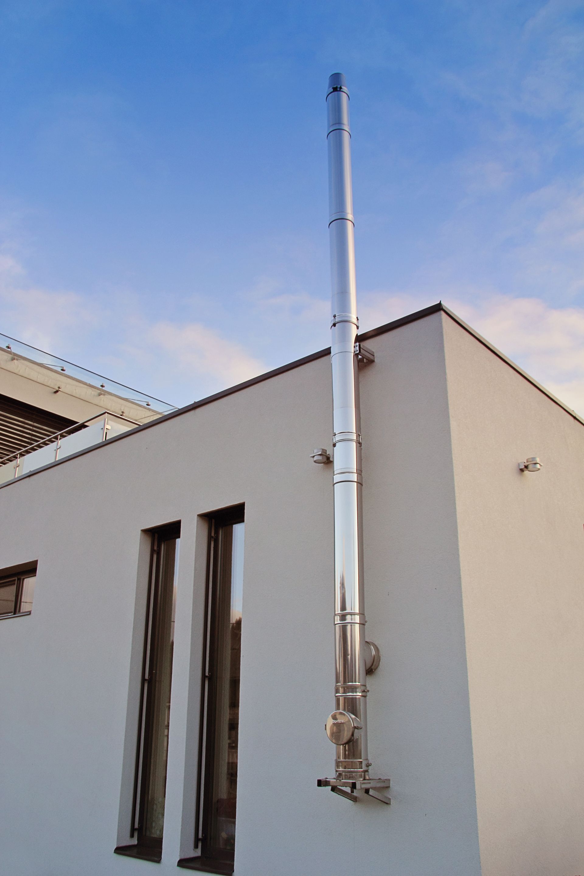 Cheminée extérieure adossée à un bâtiment blanc. Conduite métallique argentée se dressant sur un ciel bleu.
