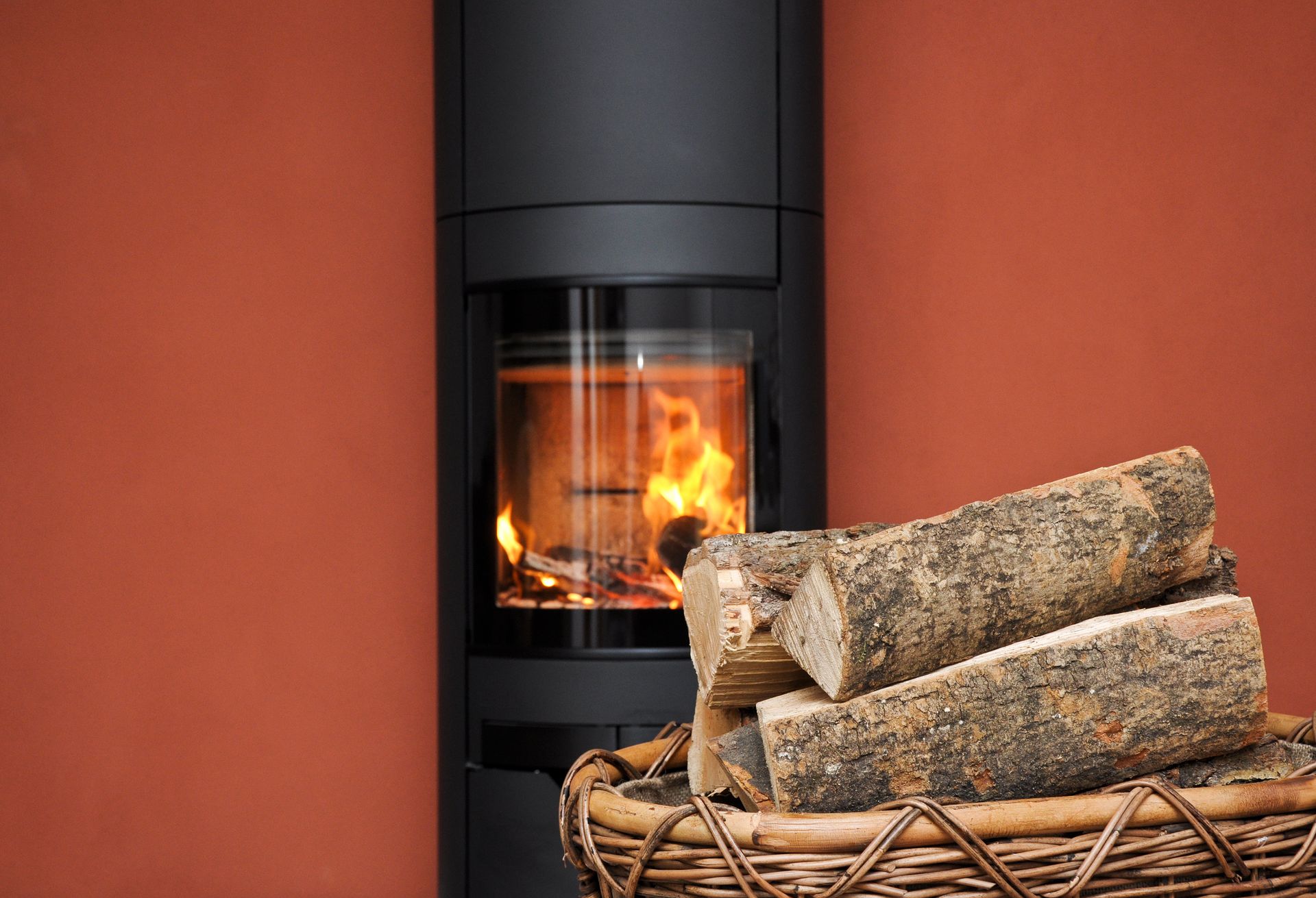 Poêle au bois avec des flammes, à côté d'un panier de bois de chauffage, contre un mur brun rougeâtre.