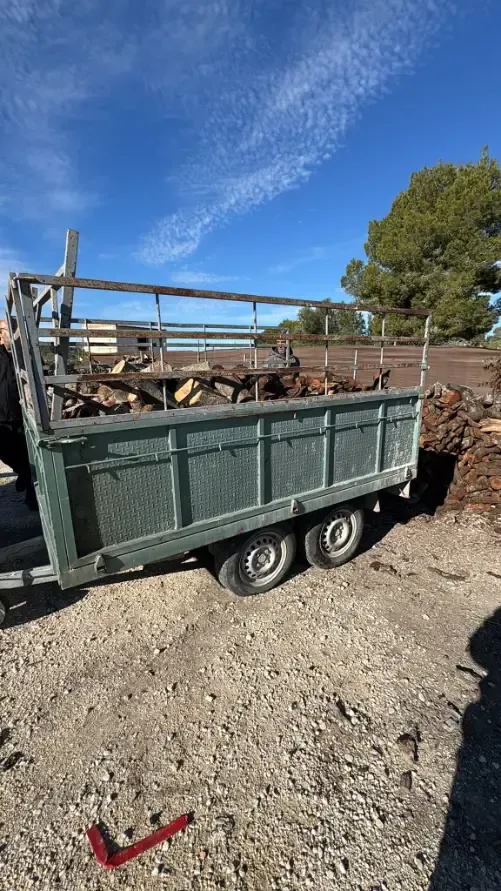 Remolque verde cargado de chatarra de madera y metal en un terreno de grava bajo un cielo azul.