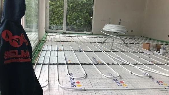 Un homme en sweat à capuche observe une installation de chauffage au sol. Tuyaux blancs sur une grille verte.