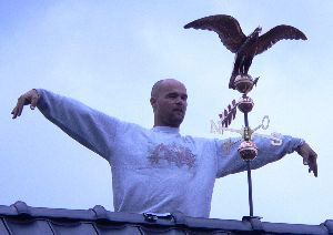 Ein Mann steht mit ausgestreckten Armen auf einem Dach und hat eine Wetterfahne mit einem Adler darauf.