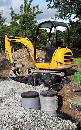 Une pelleteuse jaune sur un chantier de construction, près de gravier, d'une bâche noire et de structures circulaires grises.