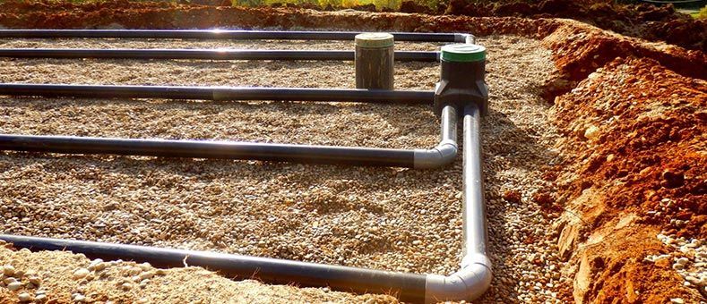 Tuyauterie et boîte de distribution du système septique enterrées dans un lit de gravier, avec terre et herbe apparentes.