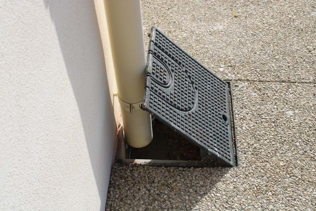 Plaque d'égout ouverte à côté d'une descente de gouttière sur une surface en béton texturé.