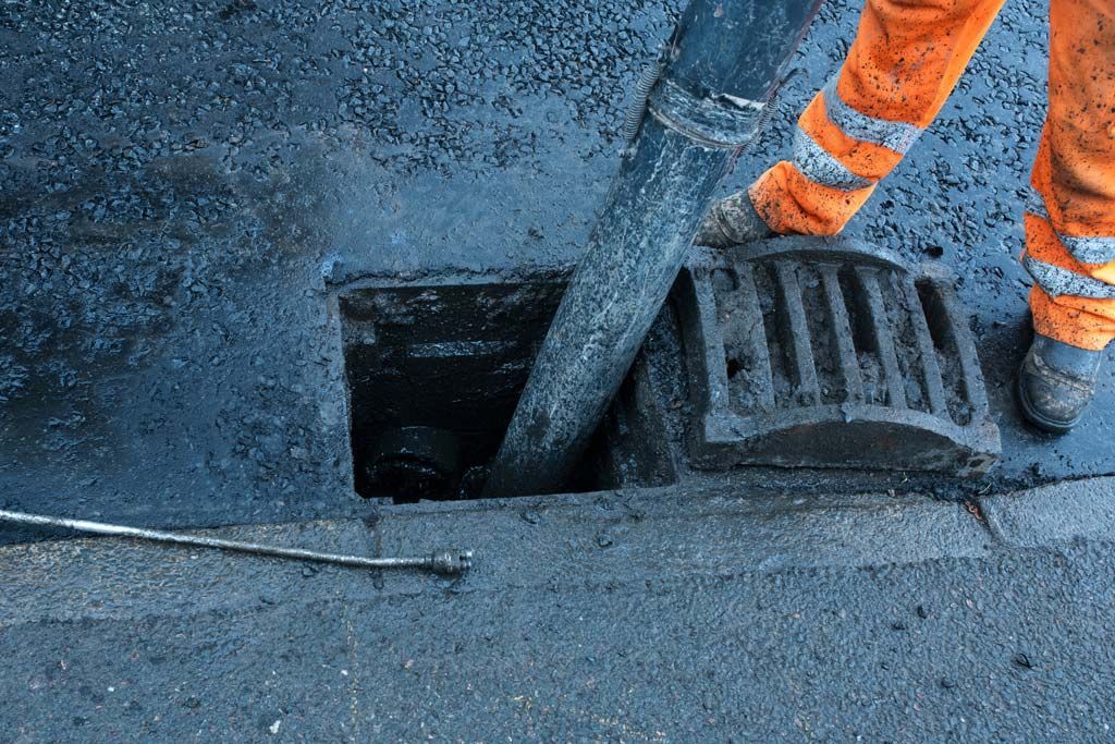 Un ouvrier en salopette orange pose de l'asphalte ; un tuyau noir se jette dans un égout.