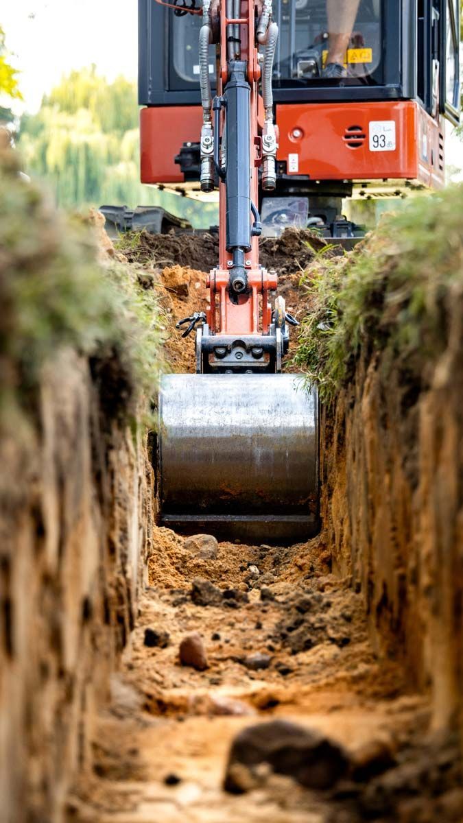 Une mini-pelle creuse une tranchée, engin orange et gris dans une zone herbeuse, soleil.