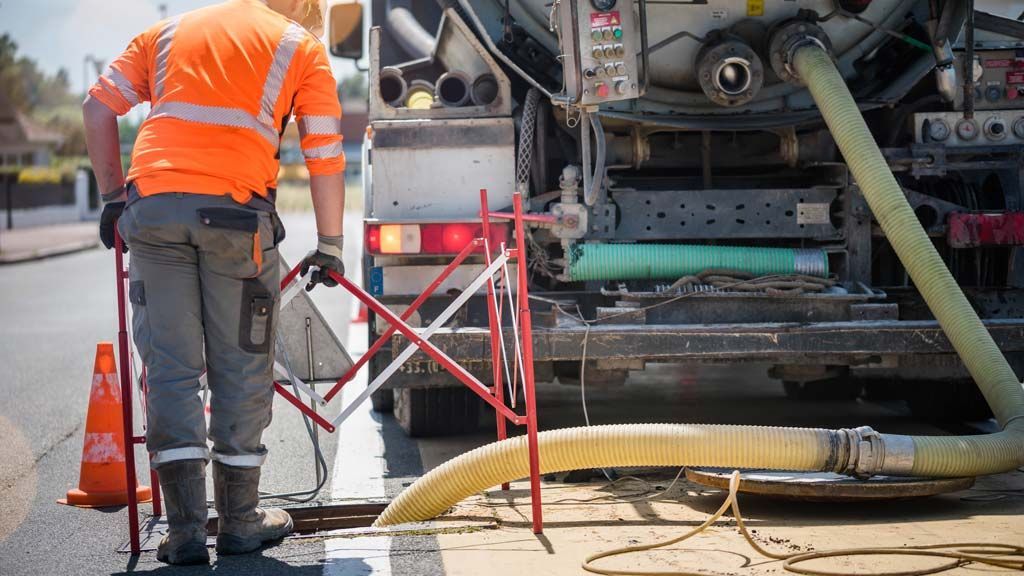 Un ouvrier du bâtiment, vêtu d'un gilet de sécurité orange, vide une canalisation d'égout à côté d'un camion de collecte des ordures.