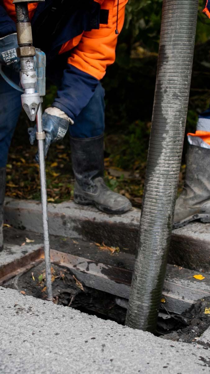 Un ouvrier utilise un outil pour travailler sur un drain en béton. Il porte une veste orange et un tuyau en caoutchouc.