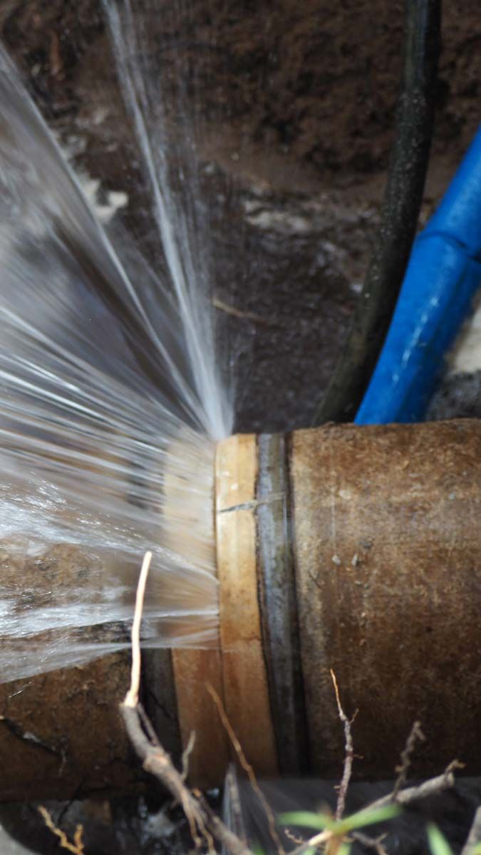 Une canalisation d'eau éclatée projette de l'eau. On aperçoit un tuyau bleu sur le tuyau marron ; l'eau jaillit avec force.