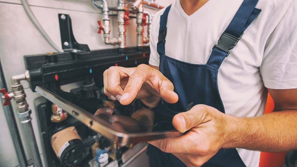 Un homme à tout faire en salopette bleue utilise une tablette près de tuyaux et de vannes industrielles.