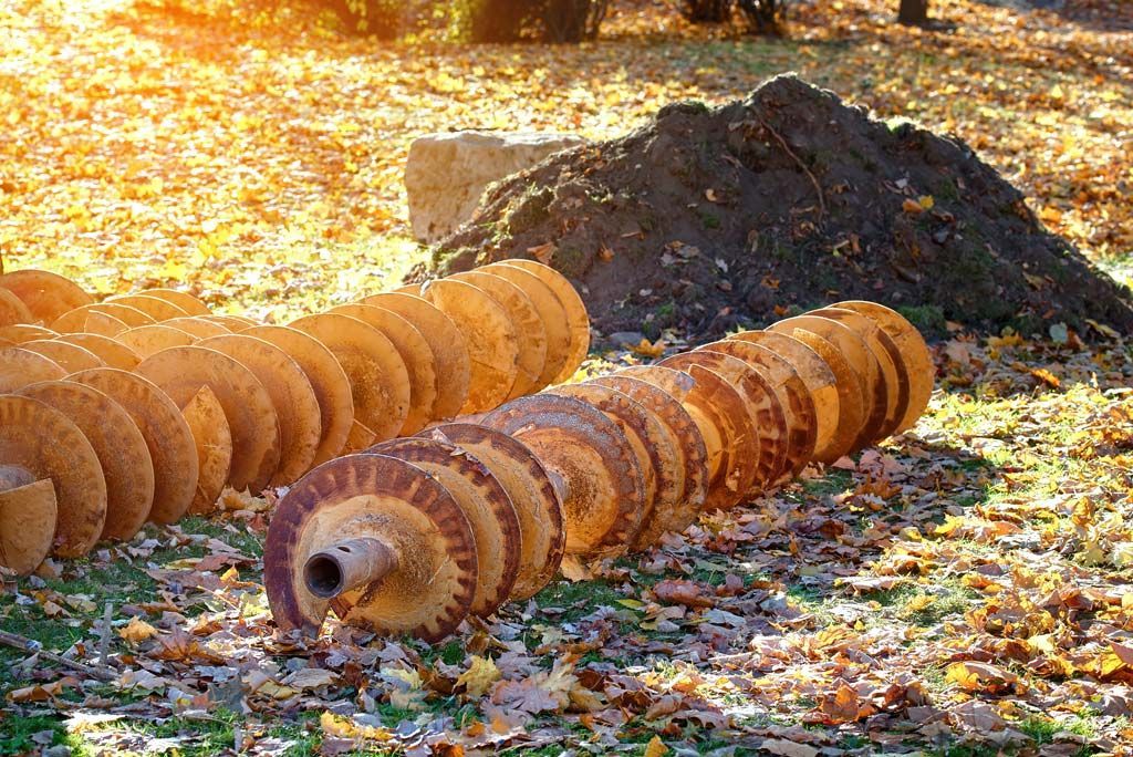 Mèches de tarière rouillées et circulaires sur un sol recouvert de feuilles mortes, près d'un tas de terre.