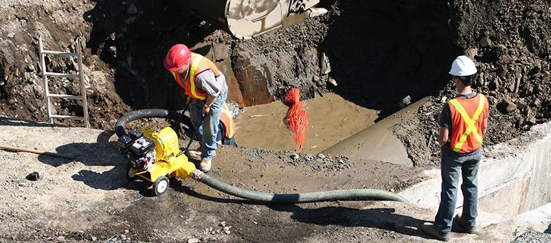 Des ouvriers portant des gilets de sécurité et des casques de chantier travaillent sur un site d'excavation où se trouvent des engins et une grosse canalisation.