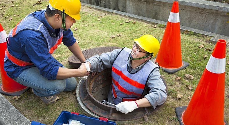 Deux ouvriers, l'un entrant dans un regard d'égout, aidé par l'autre. Tous deux portent des gilets de sécurité et des casques de chantier, entourés de cônes de signalisation orange.