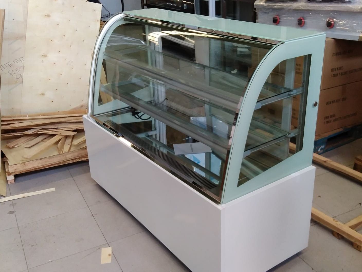 Vitrina de cristal curvada vacía en un almacén, con una base blanca y estantes apilados cerca.