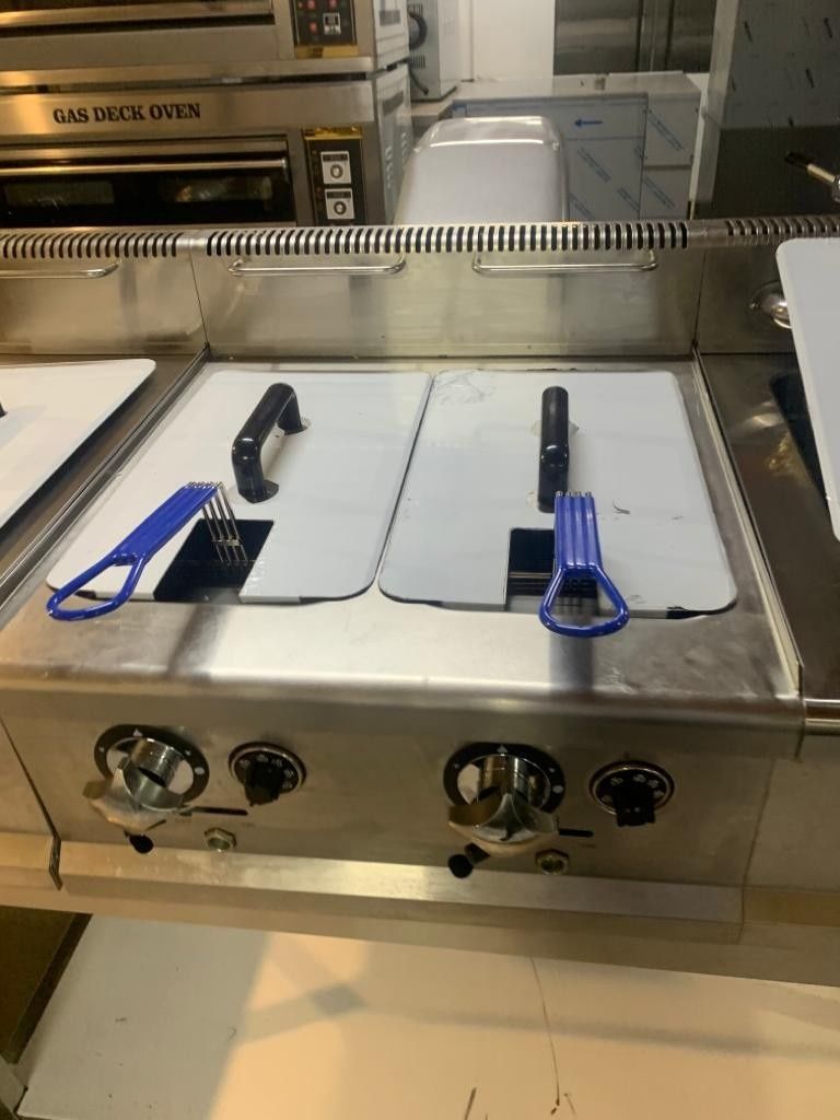Plancha comercial de acero inoxidable con dos quemadores y espátulas de mango azul sobre una encimera de cocina.
