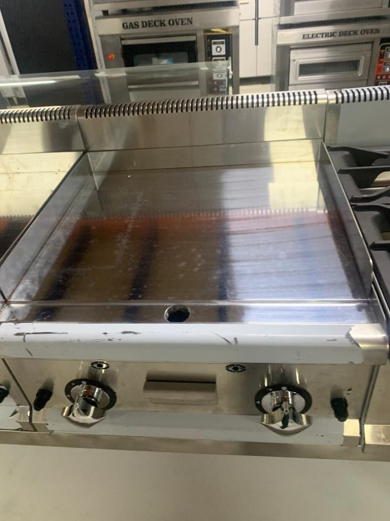Plancha o parrilla comercial de acero inoxidable con perillas de control en exhibición de electrodomésticos de cocina.