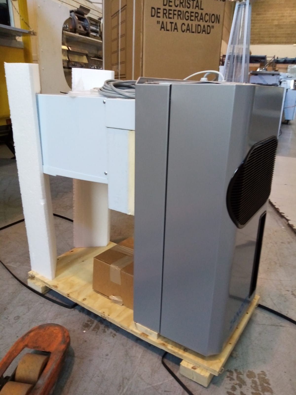 Máquina industrial con paneles grises y una rejilla de ventilación circular negra sobre un palé de madera en un taller.