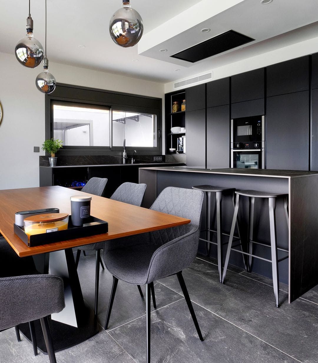 Cocina moderna con gabinetes negros, isla y mesa de comedor; sillas grises.