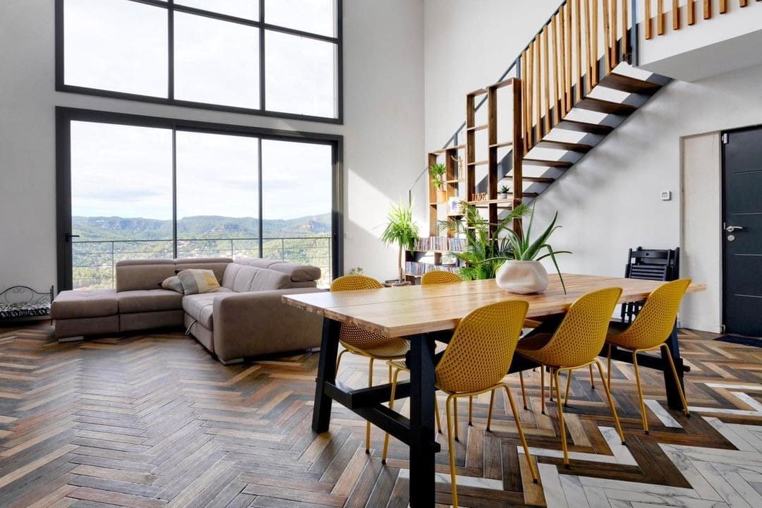 Sala de estar de planta abierta con mesa de comedor, sofá seccional, escaleras y grandes ventanales con vista al exterior.